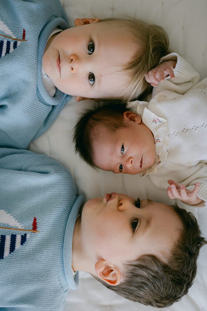 Two brothers in matching sweaters are lying on the bed with their newborn baby sister nestled between them, all three heads in row in their home in the Connecticut suburbs. 