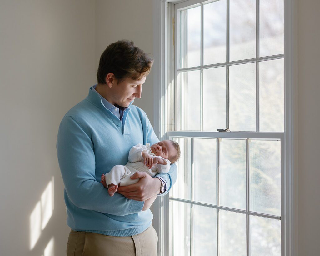 A father is holding his newborn baby girl in the crook of his arm while standing into of a sunlit window in his home in the Connecticut suburbs. 