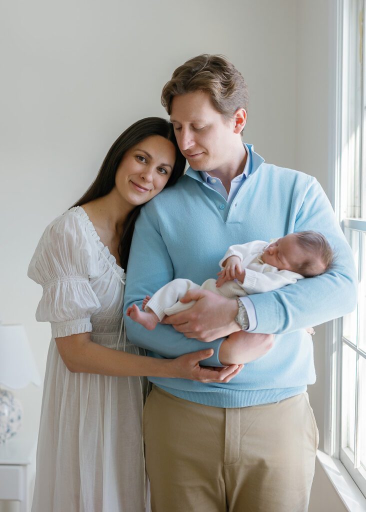 A couple stand in a loving embrace while holding their newborn daughter near the light of the window in the Connecticut suburbs.