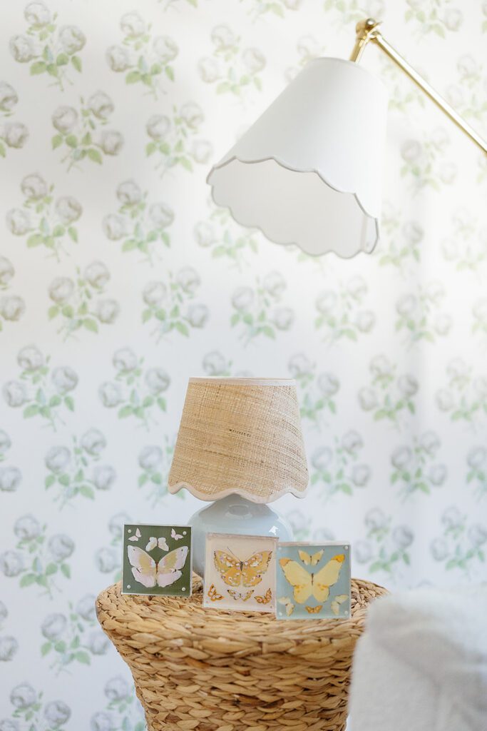 A floral wallpaper adorns the nursery of a baby girl in the suburbs of Connecticut, where three pieces of butterfly artwork sit on the seagrass side table with a scalloped floor lamp overhead. 