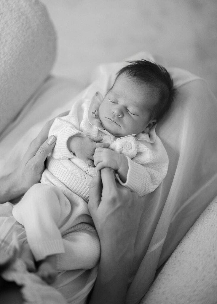 A newborn baby girl is nestled sleeping on the lap of her mother, gripping her index finger with one hand, while at home in the Connecticut suburbs. 