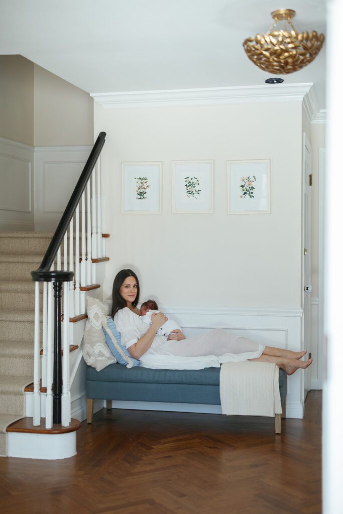 Mom is laying on an upholstered bench at the base of the stairwell in her home in the Connecticut suburbs, her newborn baby girl resting on her chest. 