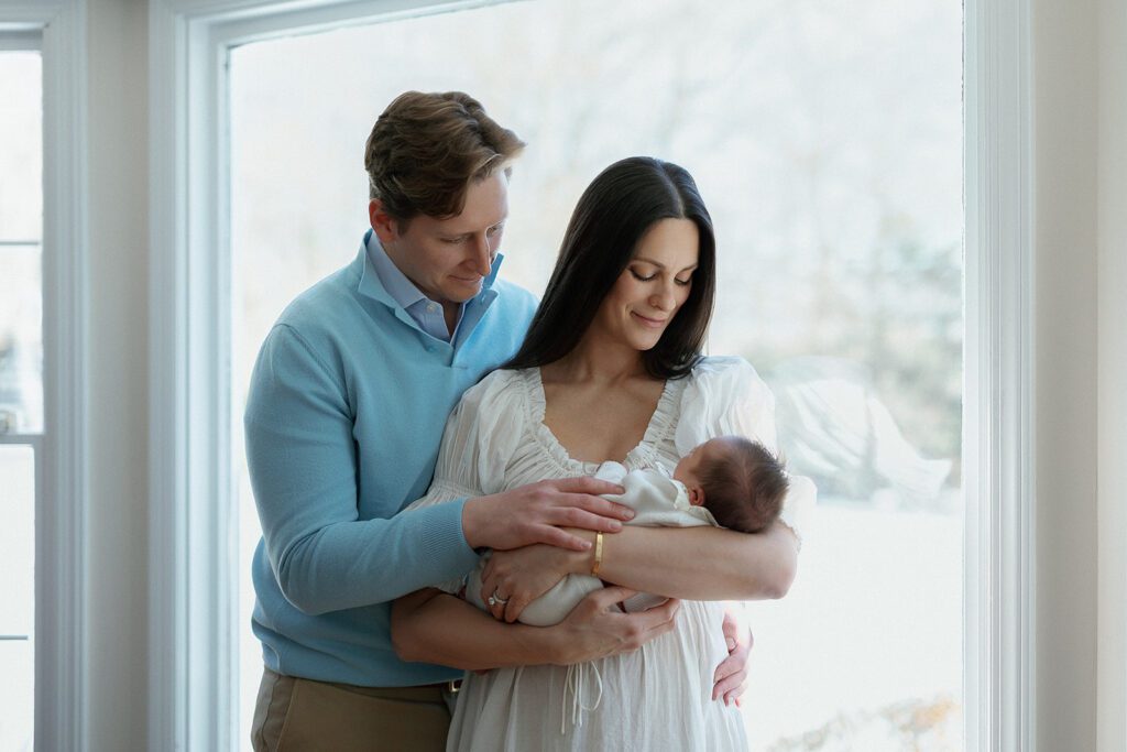 A couple stand in a loving embrace looking down at their newborn daughter in front of a large light-filled window in their home in the Connecticut suburbs.