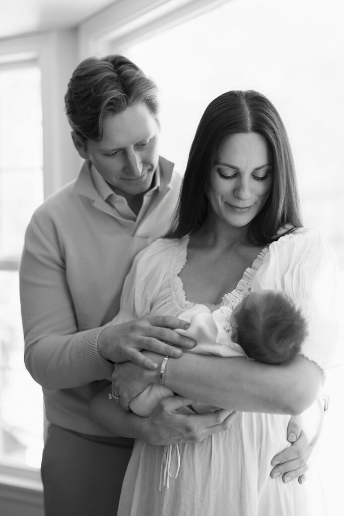A couple stand in a loving embrace looking down at their newborn daughter at home in the Connecticut suburbs.