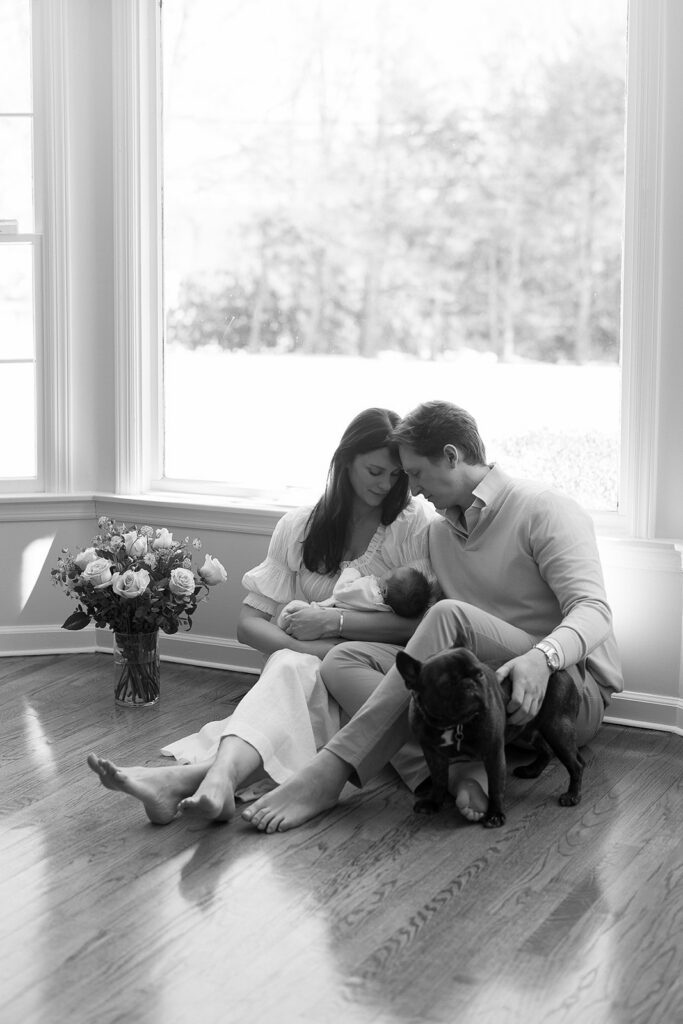 A husband and wife are sitting on the floor in front of a large window in their home in the Connecticut suburbs, looking down at their newborn baby in momma's arms, surrounded by a vase of roses and their french bulldog. 
